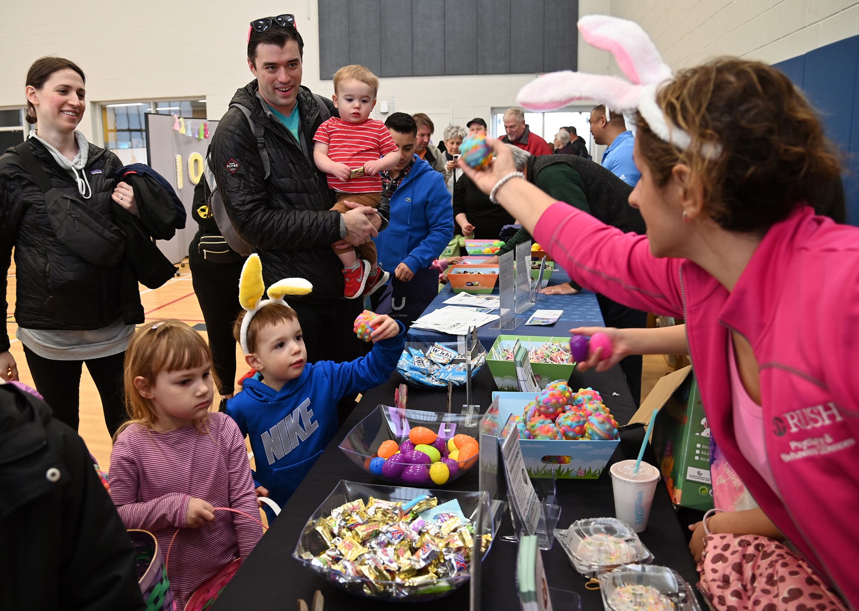 Top left, the Sturgeon family of Mundelein collect eggs and treats along the path. The parents are Andy and Samantha Sturgeon with their children Kai, 1, and Jay, 3. On right, handing them an egg is sponsor table representative Nicole Russo-Ponsaran of Mundelein and of RUSH NeuroBehavioral Center at the Cottontail Trail event on March 16, 2024 at the Dunbar Recreation Center at Community Park in Mundelein. (Karie Angell Luc/Lake County News-Sun)